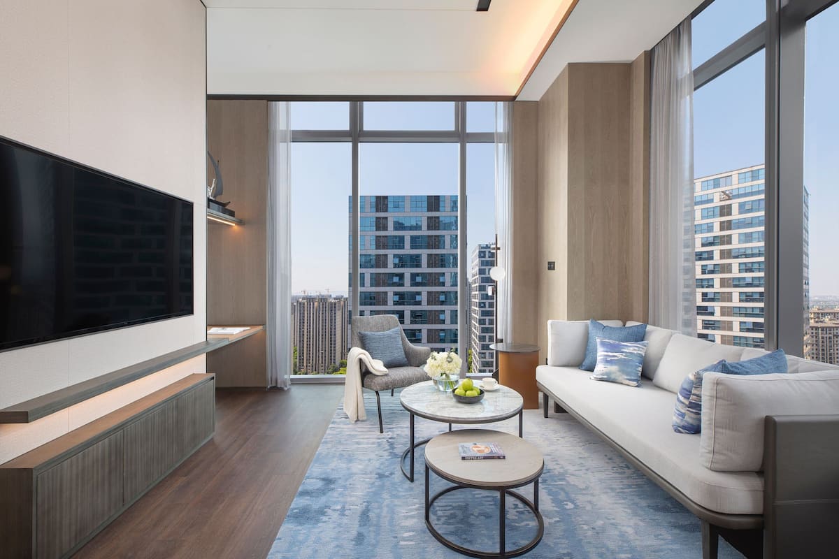 A modern living room with a large flat-screen TV, a sofa, a coffee table, and a chair. There is a large window with a view of high-rise buildings.