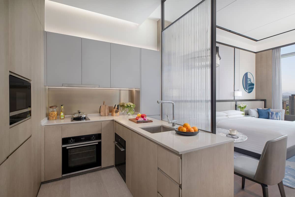 A modern hotel room with a kitchen area, a bed, and a large window.