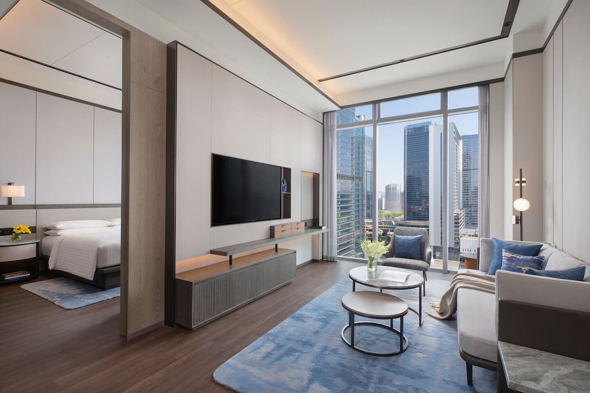 A modern hotel room with a large TV, a sofa, a coffee table, and a view of the city skyline.