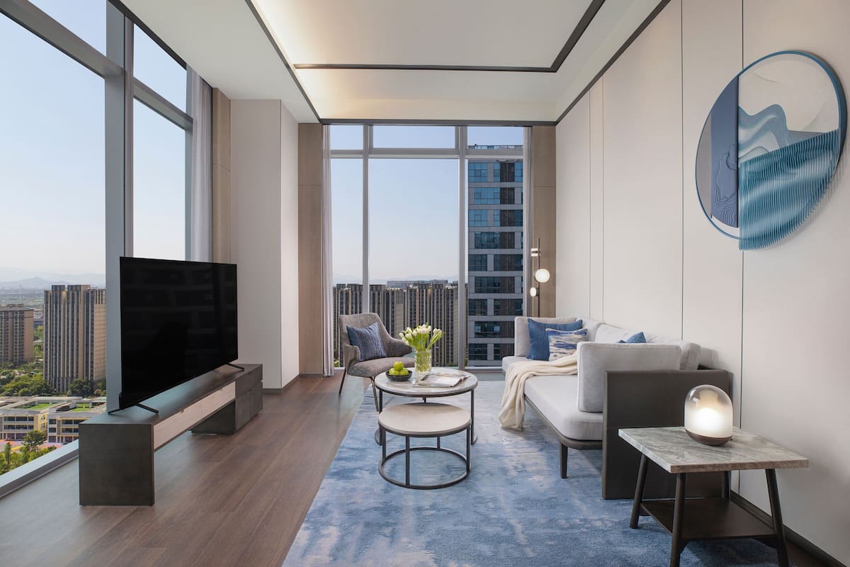 A modern living room with a large TV, comfortable seating, a coffee table, and a view of the cityscape through large windows.