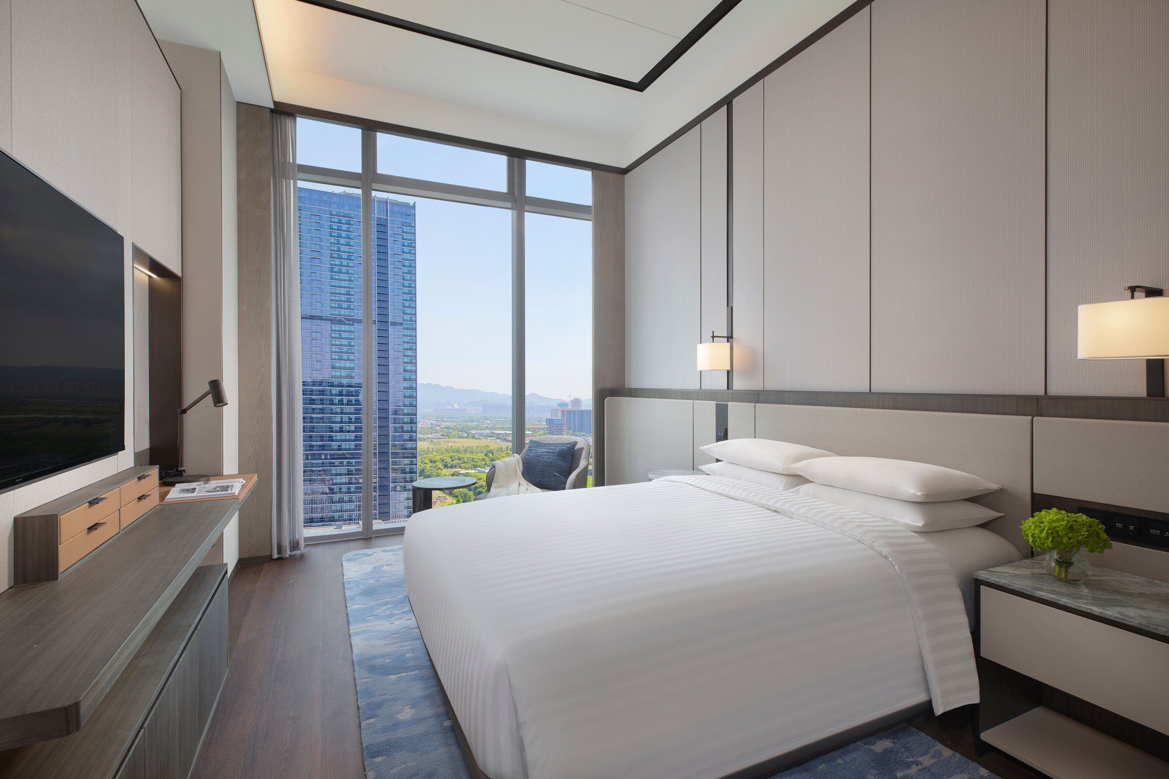 A modern hotel room with a large bed, a flat-screen TV, a desk, and a view of a cityscape through large windows.