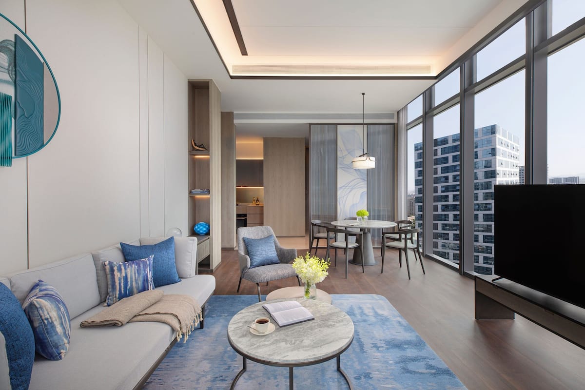 A modern living room with a sofa, coffee table, and dining area. There is a large window with a city view.