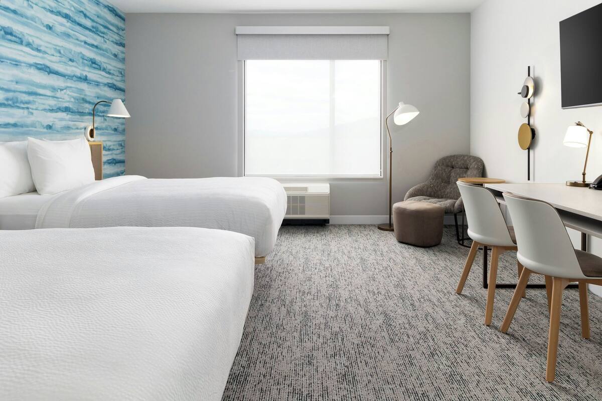Et hotelværelse med to senge, et skrivebord med stole, et stort vindue med persienner, et vægmonteret fjernsyn og et tæppe med mønster.