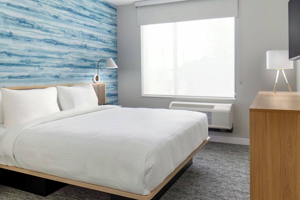 Et hotelværelse med en seng, et natbord, en lampe, et vindue med udsigt og en aircondition.
