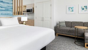 A hotel room with a large bed, a gray sofa, a small round table, and a microwave in the kitchenette.