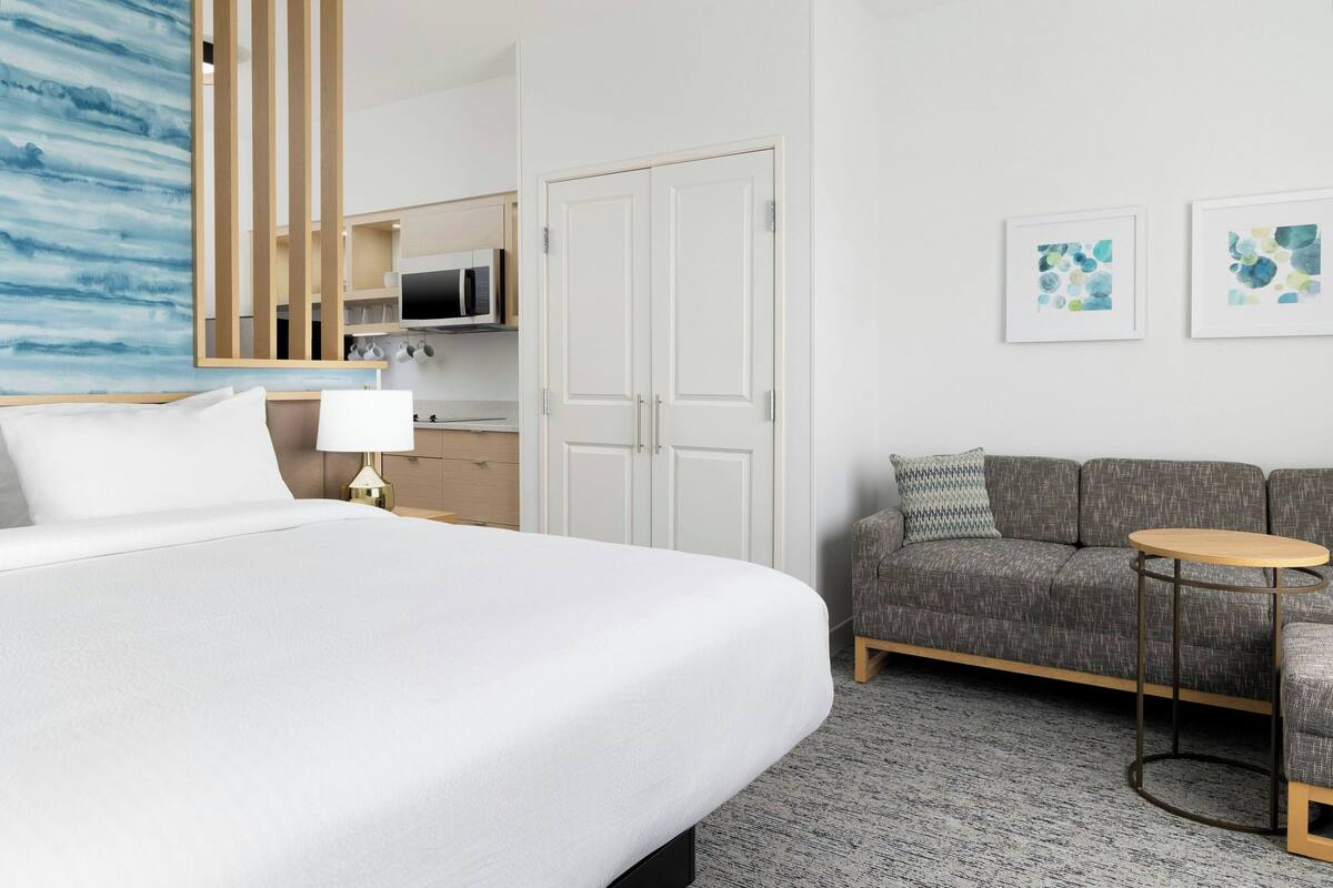 Et hotelværelse med en stor seng, en grå sofa, et lille rundt bord og en mikroovn i tekøkkenet.