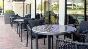 An outdoor patio with round tables and wicker chairs, a brick floor, and a view of the street through large windows.