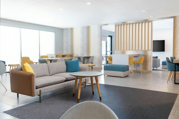 Un hall d’hôtel moderne avec un canapé, des fauteuils, une petite table et un téléviseur fixé au mur.