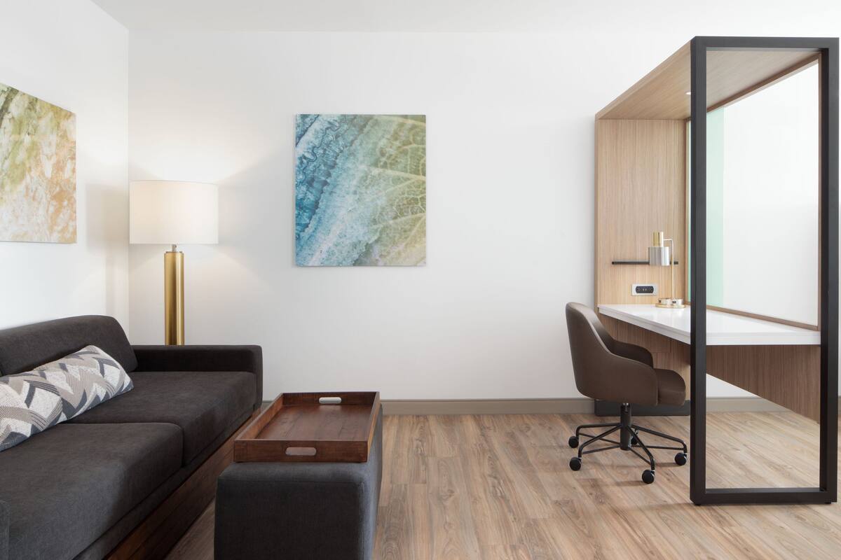 Ein modernes Hotelzimmer mit grauem Sofa, einem Holznachtkästchen, einem Schreibtisch mit Stuhl und einem großen Spiegel.