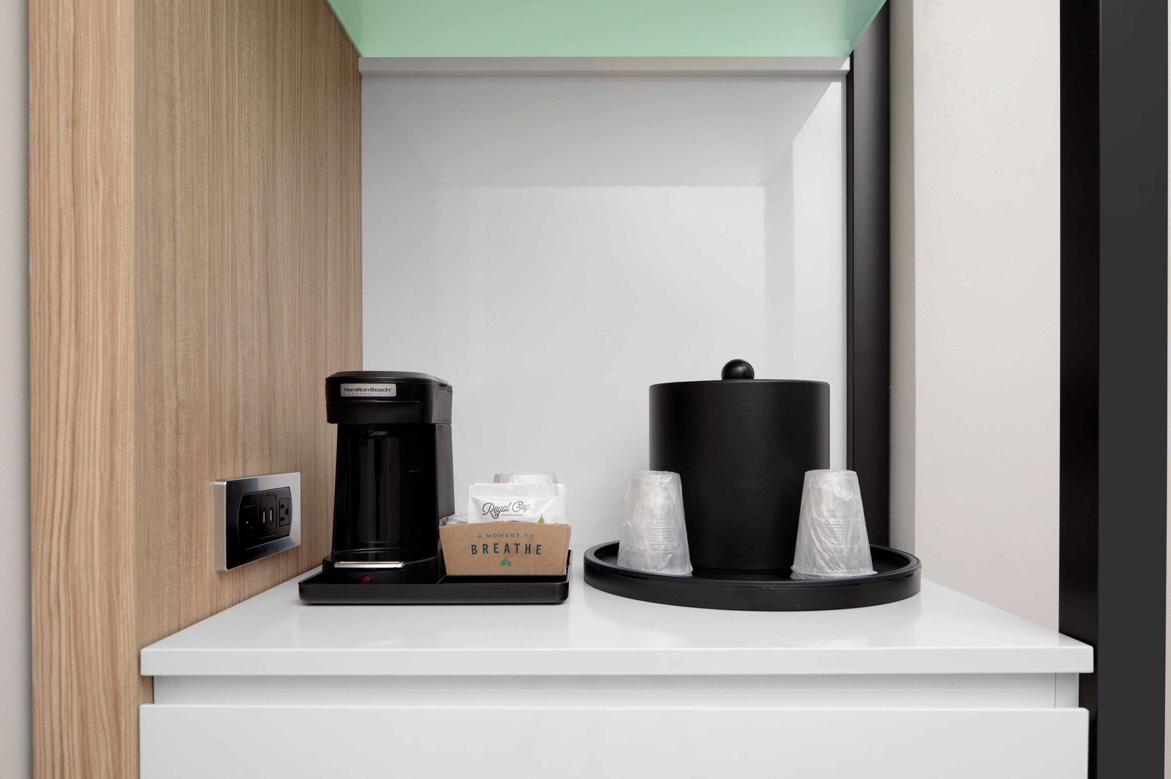 A coffee station with a coffee maker, a box labeled "A MOMENT TO BREATHE," and two cups on a tray.