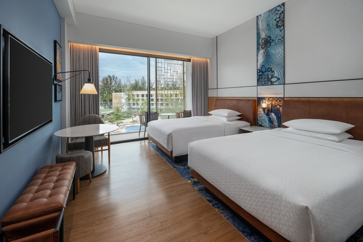 A hotel room with two beds, a desk, a bench, and a large window with a view of buildings and greenery.