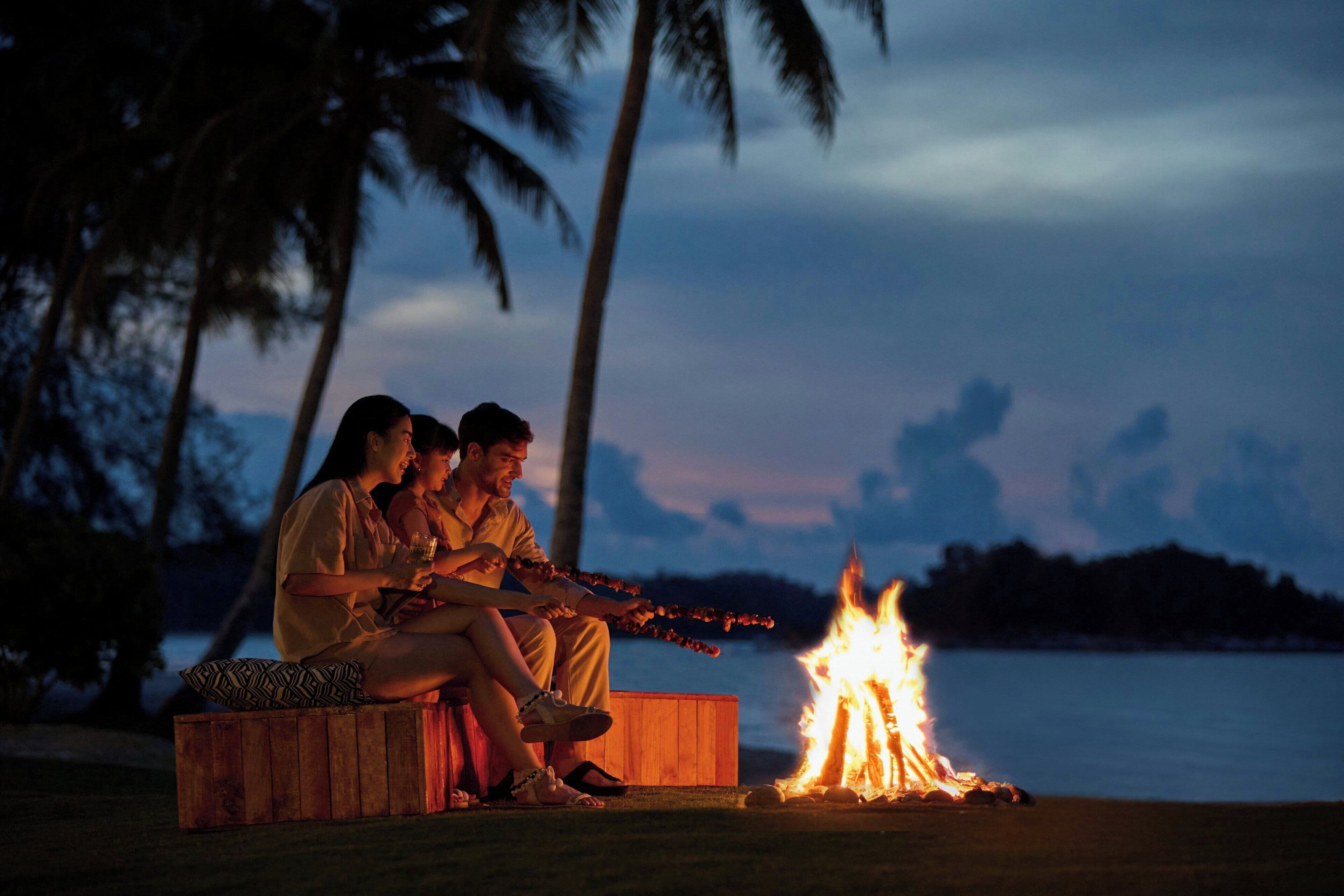 Three people sitting by a campfire on a wooden bench with a lake and palm trees in the background.