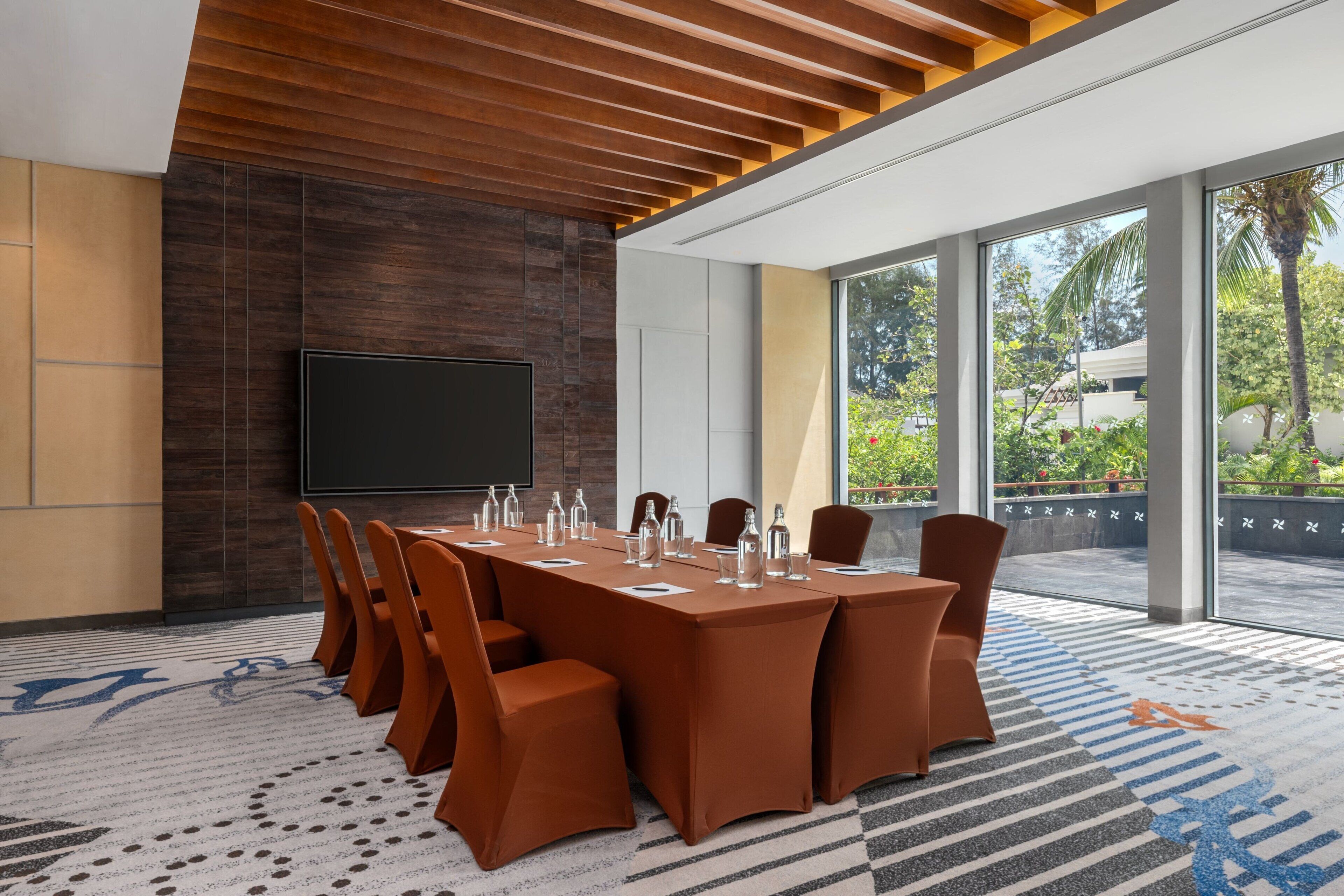 A conference room with a long table, chairs, a flat-screen TV, and large windows with a view of greenery.