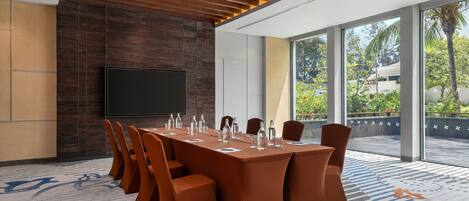 A conference room with a long table, chairs, a flat-screen TV, and large windows with a view of greenery.