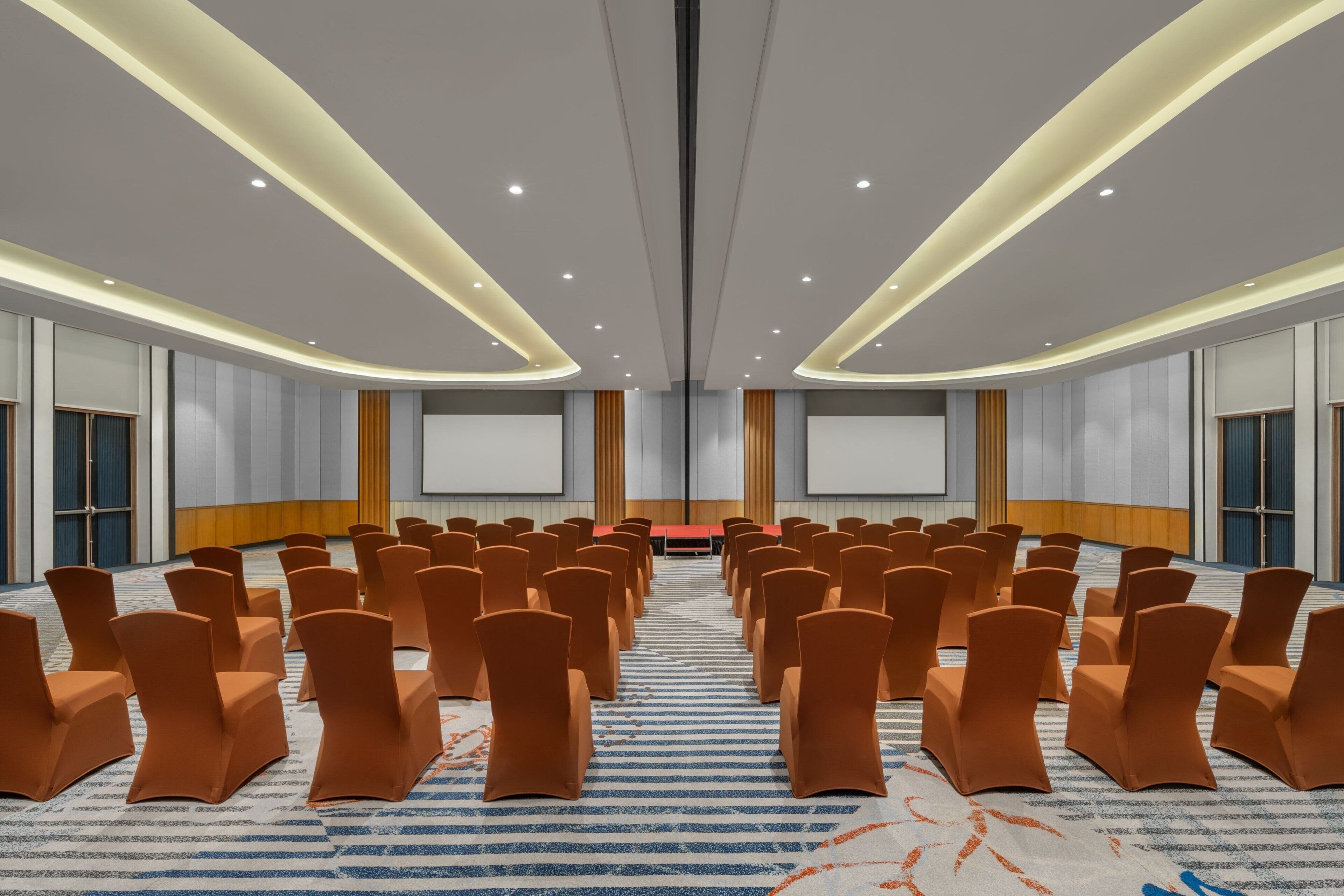 A conference room with rows of orange chairs, a red table, and a large screen at the front.