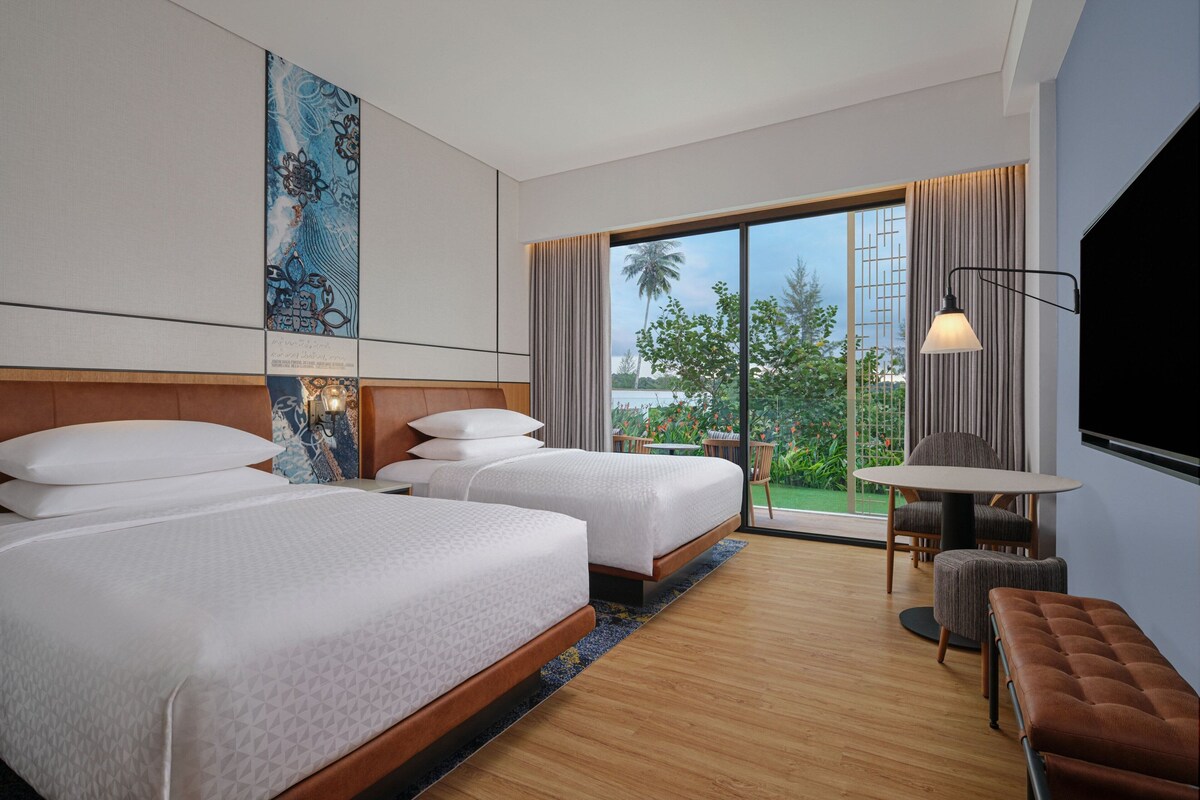 A hotel room with two beds, a TV, a table with chairs, and a balcony with a view of palm trees.