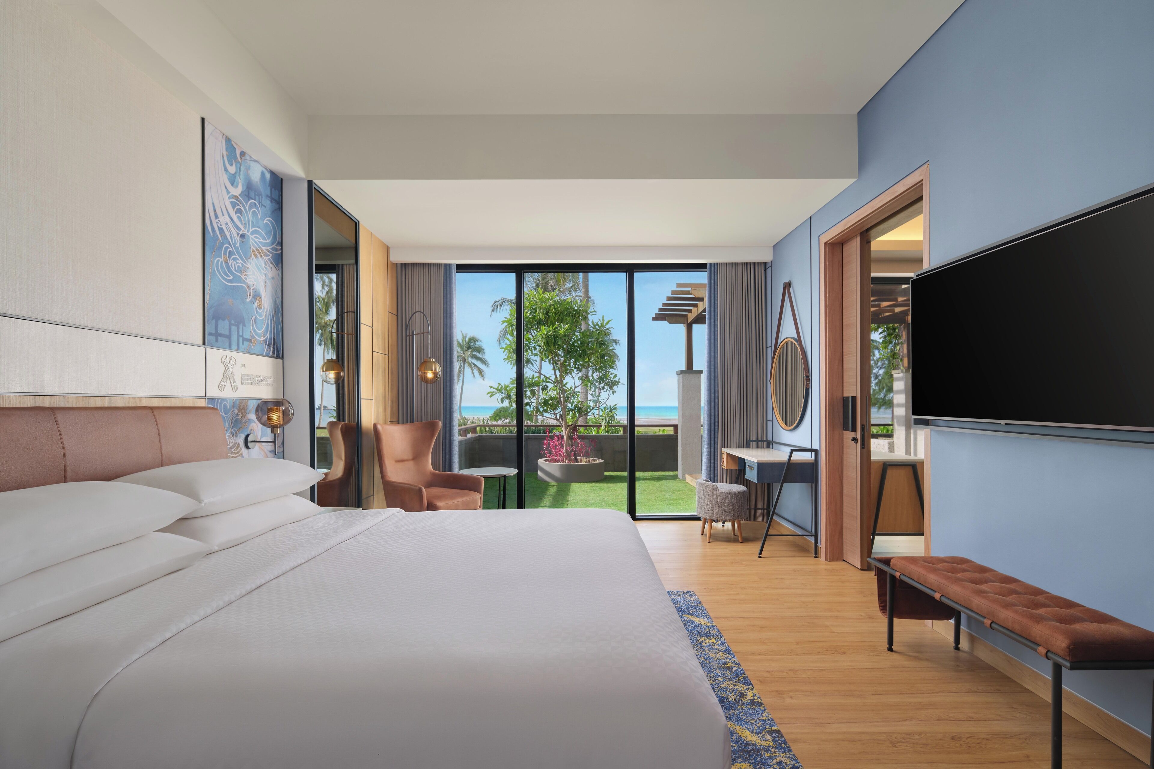 Quarto de hotel moderno com uma cama grande, televisão de tela plana, área de estar com um banco e vista para o exterior.