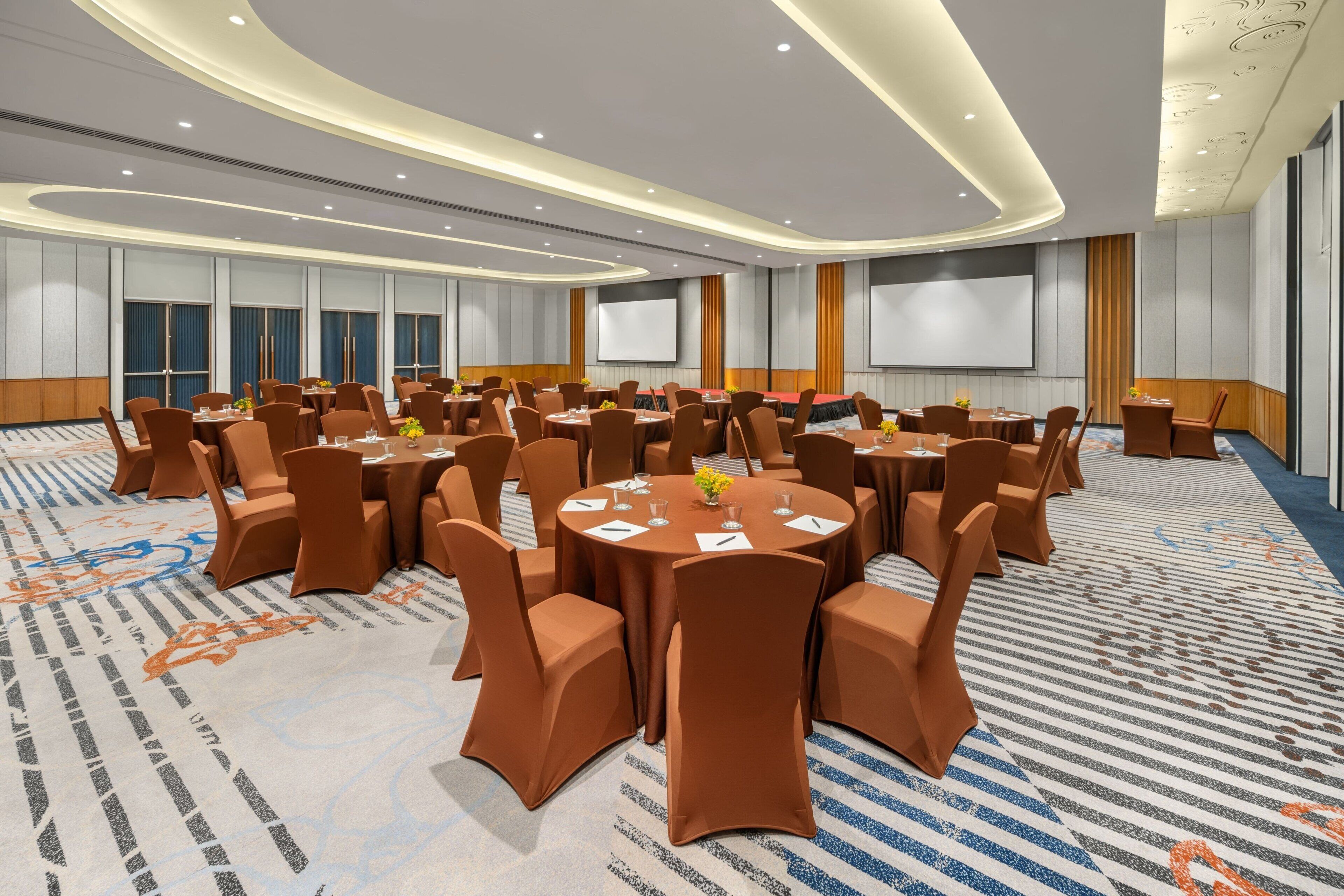 A conference room with round tables and brown chairs, equipped with screens and a carpeted floor.