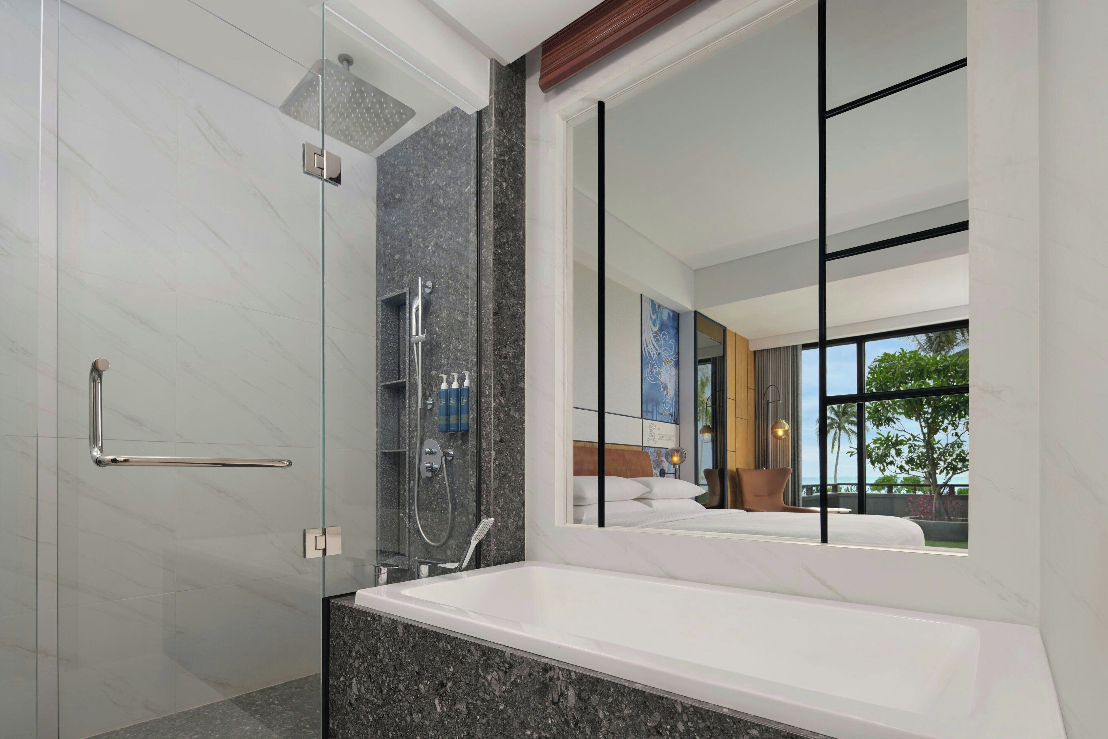 Ein modernes Badezimmer mit einer ein Glasfach stammenden Dusche, einer großzügigen Badewanne und einem großen Spiegel, der ein Schlafzimmer mit Bett und Blick ins Freie reflektiert.