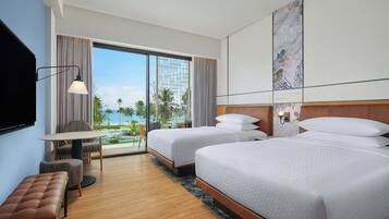A hotel room with two beds, a desk, a chair, a TV, and a view of the ocean.