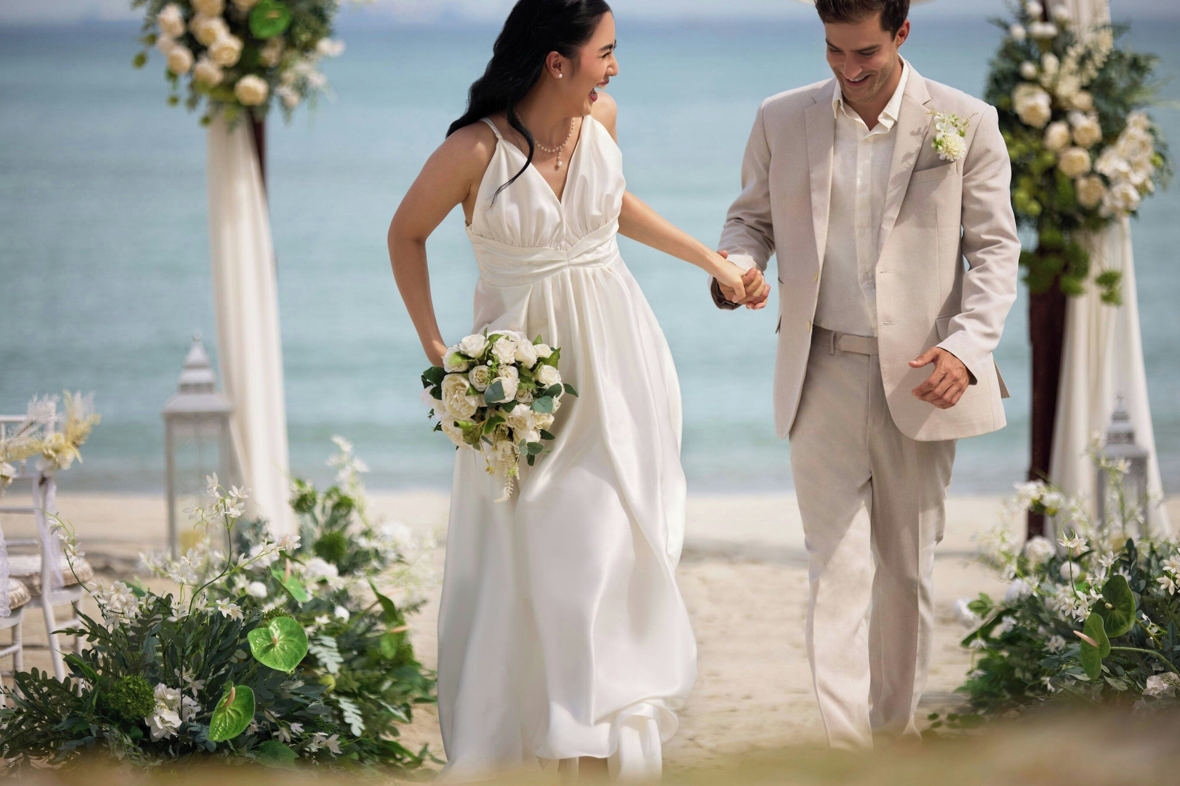 A couple walking hand in hand on a beach with floral decorations and a clear view of the ocean.
