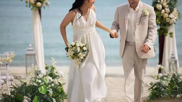 A couple walking hand in hand on a beach with floral decorations and a clear view of the ocean.