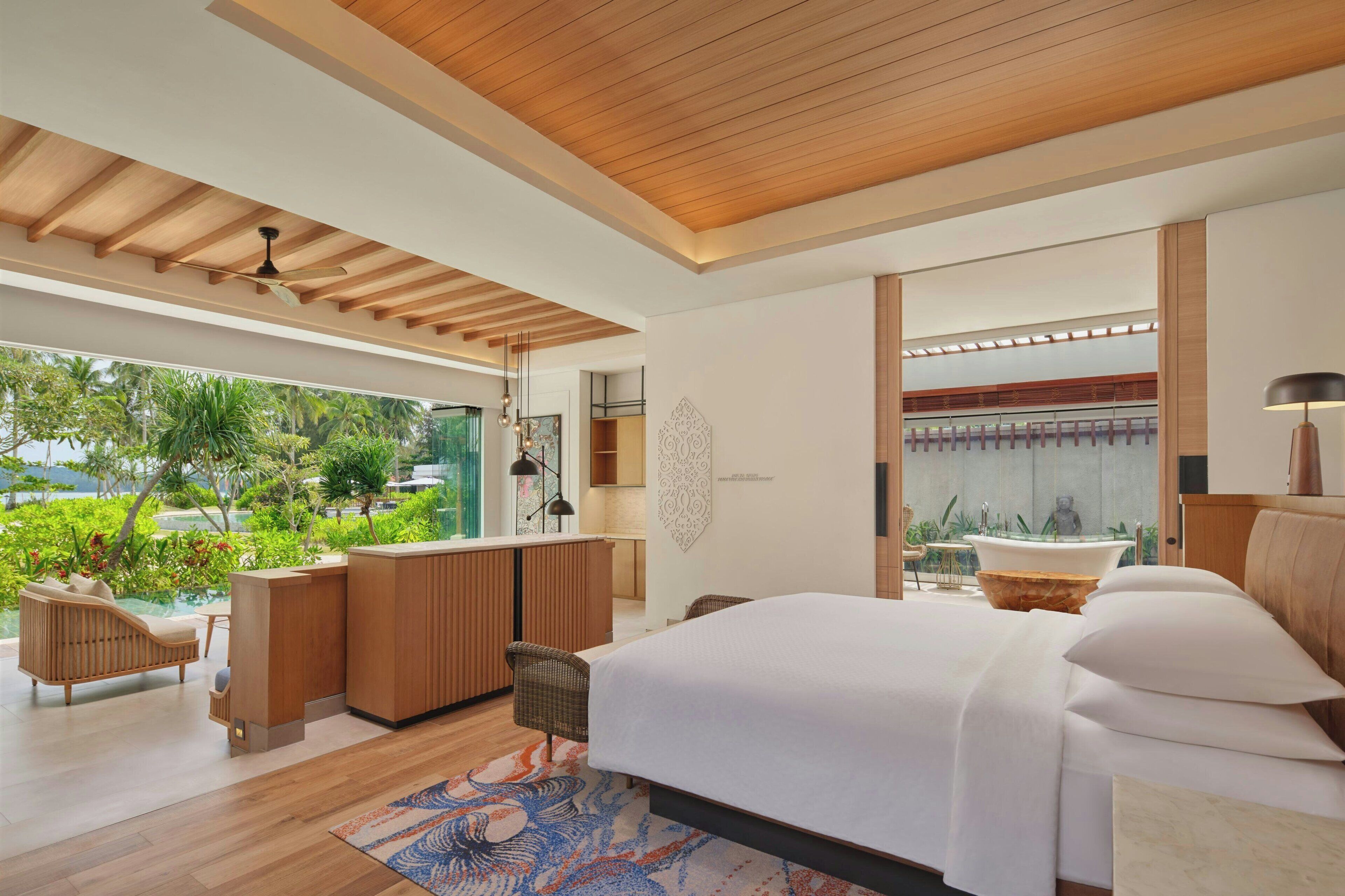 A modern bedroom with a large bed, a sitting area with a sofa and chairs, a small kitchen area, and a view of lush greenery outside.