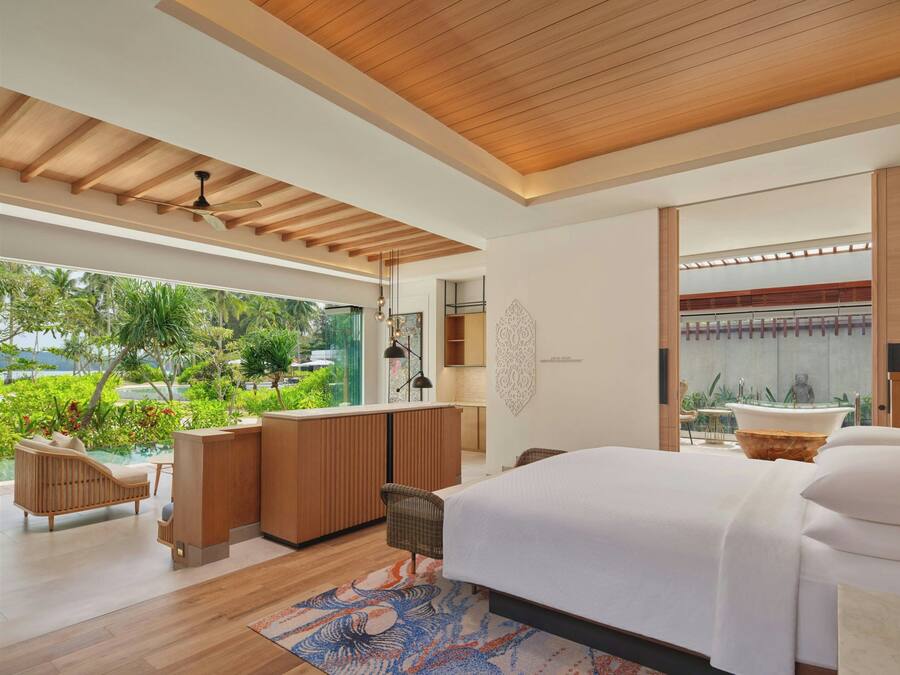 A modern bedroom with a large bed, a sitting area with a sofa and chairs, a small kitchen area, and a view of lush greenery outside.