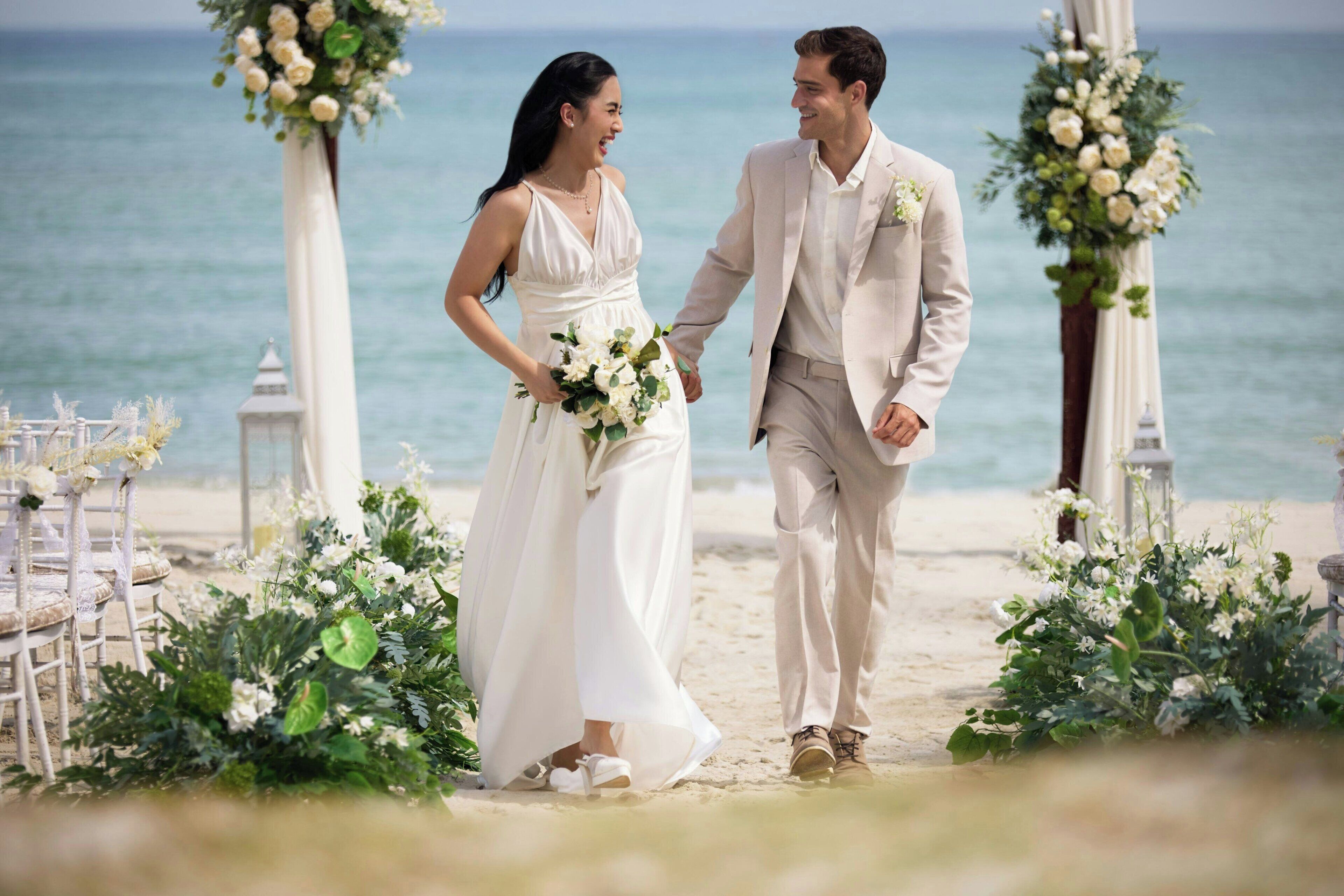 A couple walking on a beach with floral decorations and white chairs.