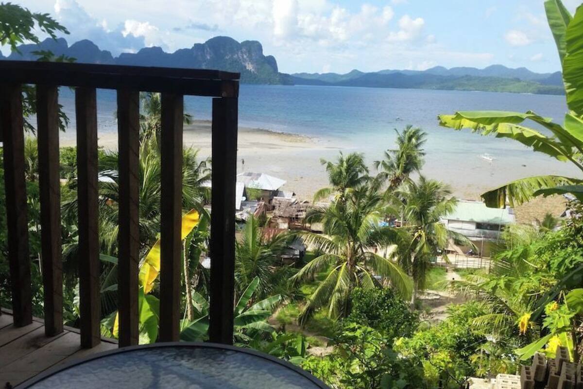 Terraza de madera con vista a una playa, palmeras y una vista del mar y las montañas.