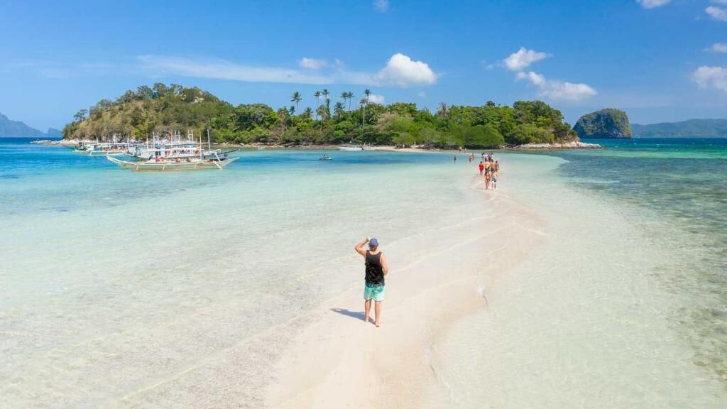 A beach with clear turquoise water, a sandy shore, and a small island with boats in the background.