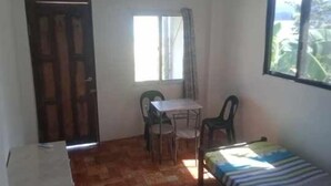 Una habitación pequeña con una cama individual, una mesa con sillas, un ventanal con cortinas y una puerta.