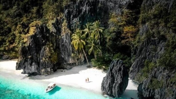 Una playa con agua turquesa cristalina, arena blanca y acantilados rocosos. Hay palmeras y un bote en la orilla.