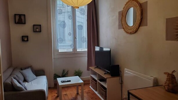 A living room with a sofa, a TV on a stand, a small table, and a window with curtains. - Hyper Centre-ville Avec Balcon, Idéalement Situé (Saint-Étienne)