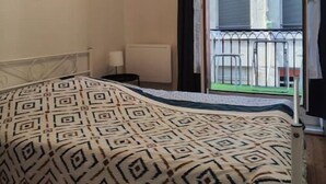 A bedroom with a patterned bedspread, a hanging light fixture, and a view of a balcony with a railing. - Hyper Centre-ville Avec Balcon, Idéalement Situé (Saint-Étienne)