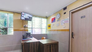 A reception area with a wooden desk, a computer, and a wall-mounted TV. There is a clock and a door with the number 108 on it. - Fabexpress Victory (Lucknow)