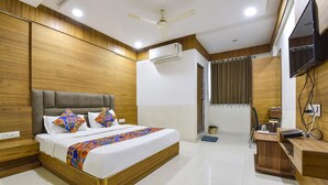 A hotel room with a wooden headboard bed, a ceiling fan, an air conditioning unit, a small desk with a chair, and a television mounted on the wall. - Fabhotel Maan Palace (Ahmedabad)