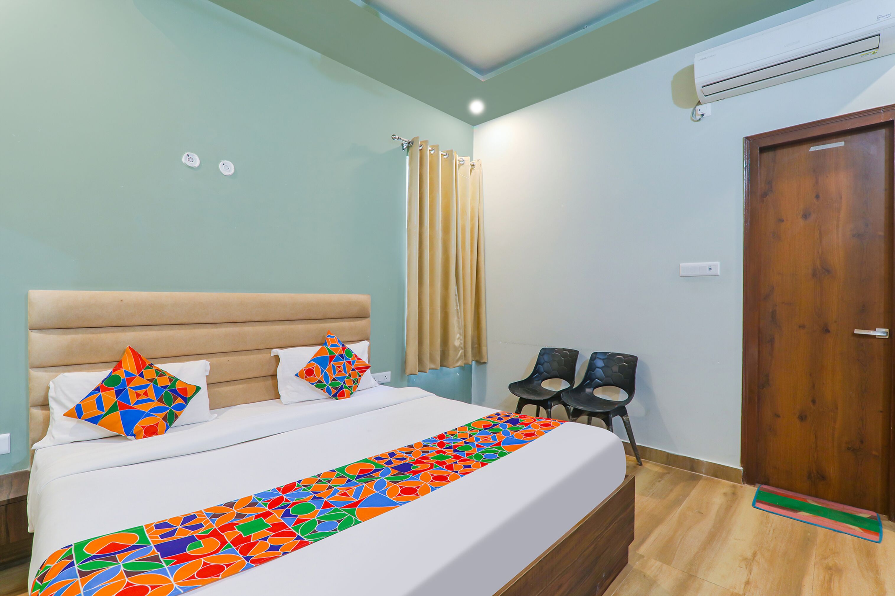 Uma cama de casal bem arrumada com um cobreleiro colorido, duas almofadas e uma cabeceira. Há duas cadeiras e uma porta de madeira no quarto.