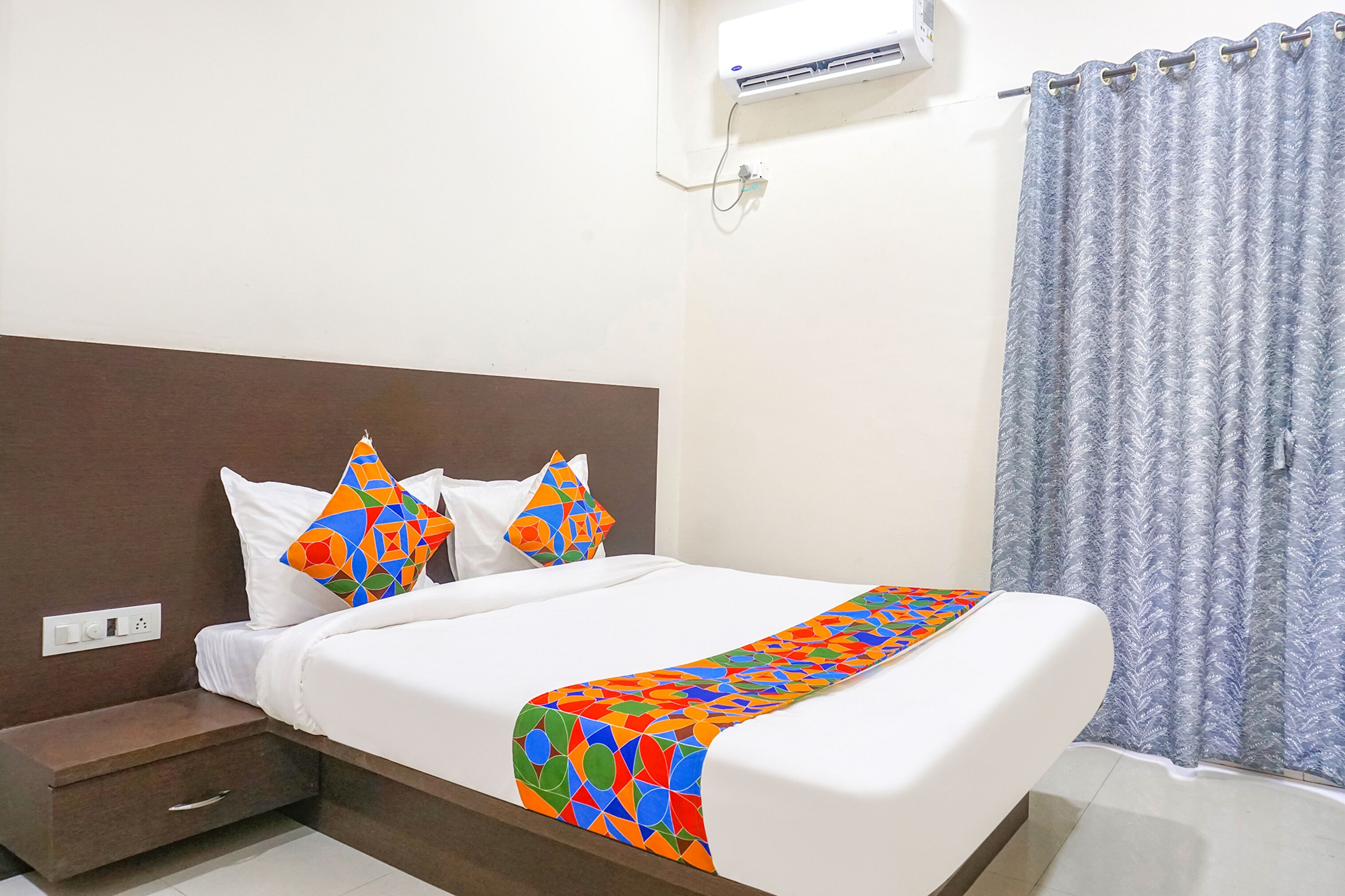 A neatly made bed with a colorful geometric-patterned bedspread, a brown headboard, and a white wall air conditioner.
