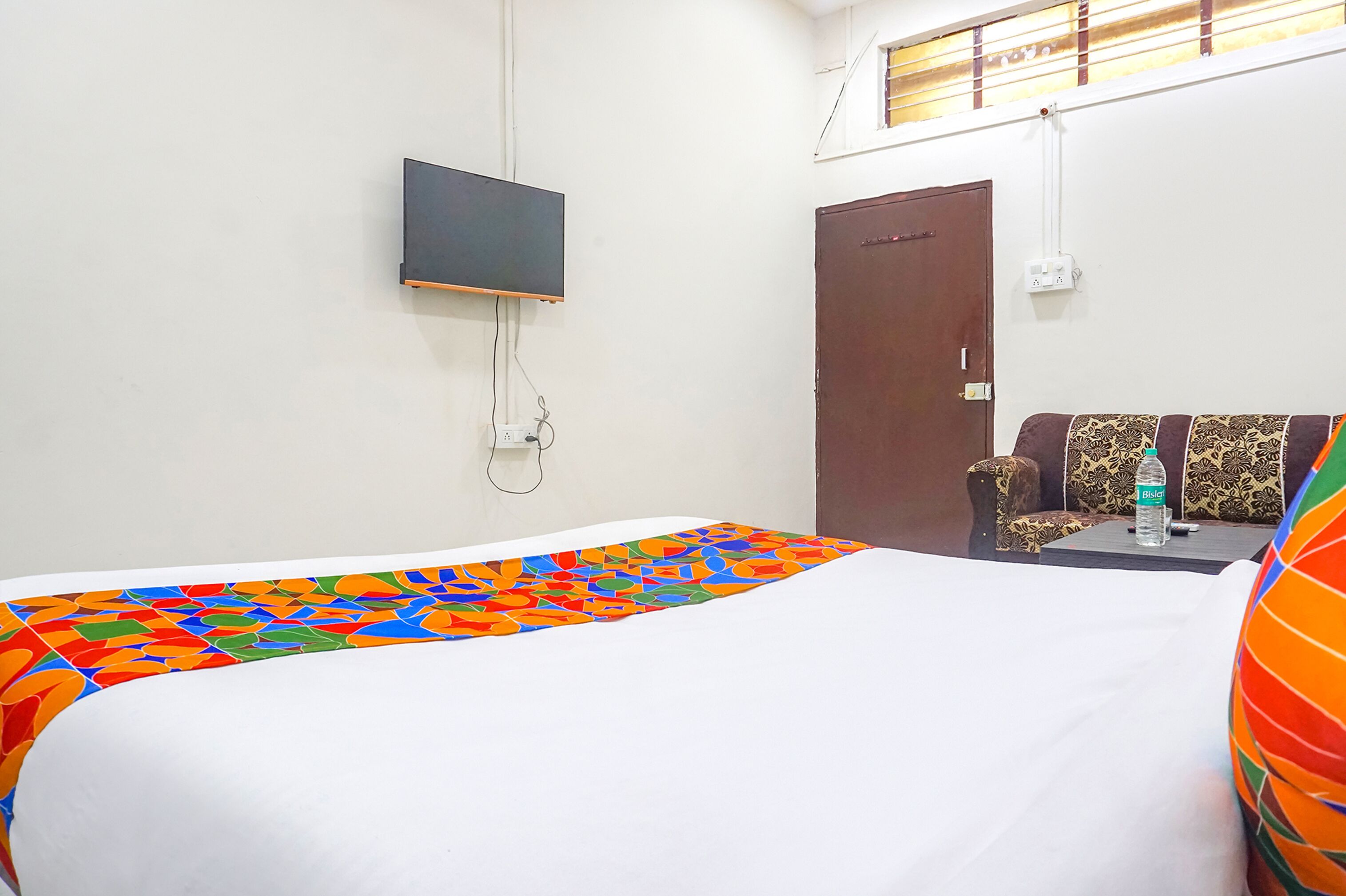 A hotel room with a bed, a television mounted on the wall, a wardrobe, and a sofa with patterned cushions.