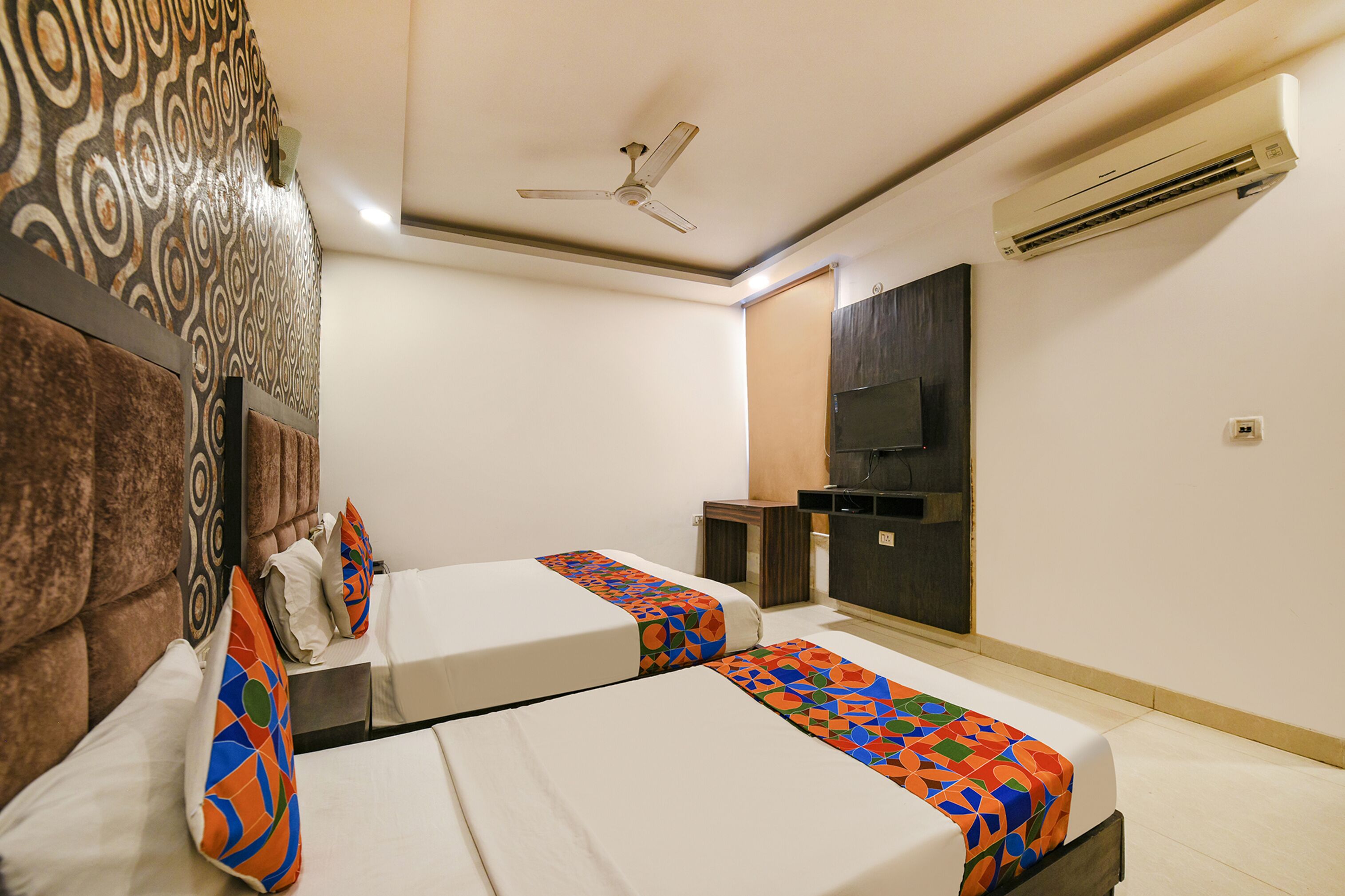 A hotel room with two beds, a ceiling fan, an air conditioner, and a small desk with a TV.