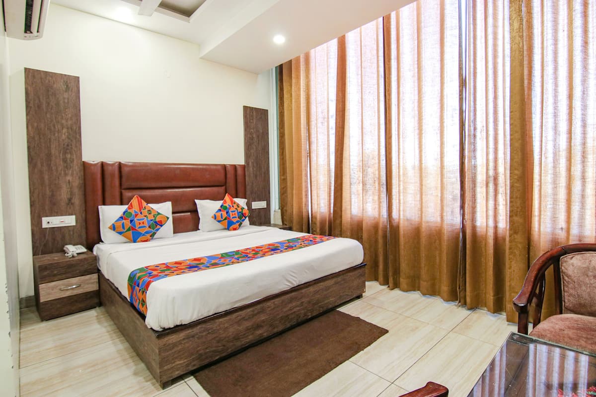 A neatly made bed with a patterned bedspread, a wooden nightstand with a phone, and a chair in a room with a large window and curtains.