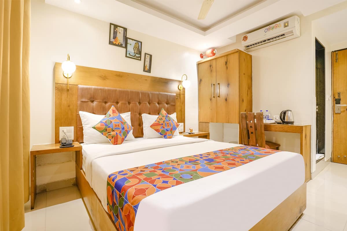 A hotel room with a large bed, wooden headboard, two bedside tables with lamps, a wardrobe, and an air conditioning unit.