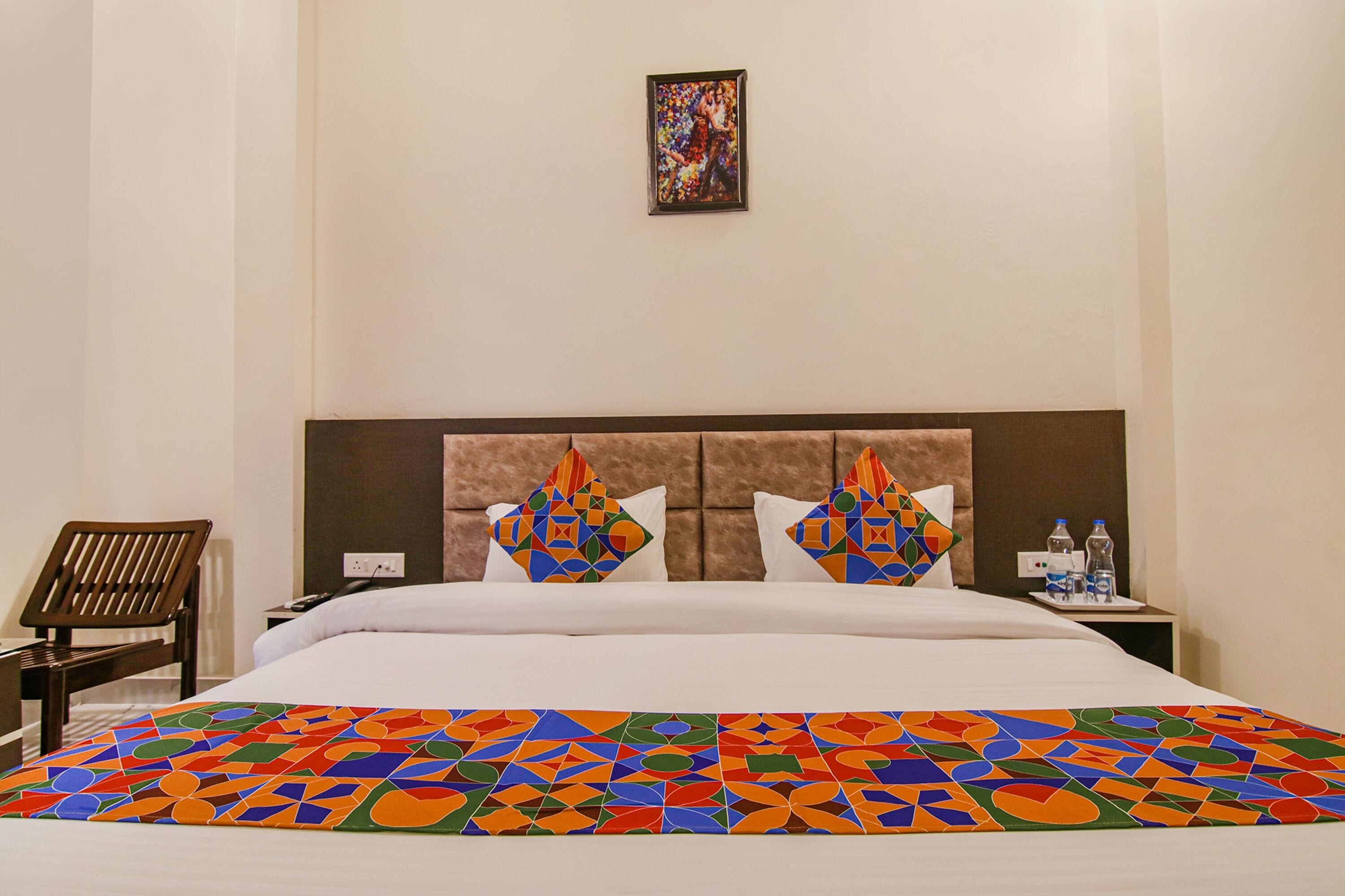 Habitación de hotel con una cama grande, cabecera con diseños geométricos, una mesita con un juego de botellas de agua y una silla.