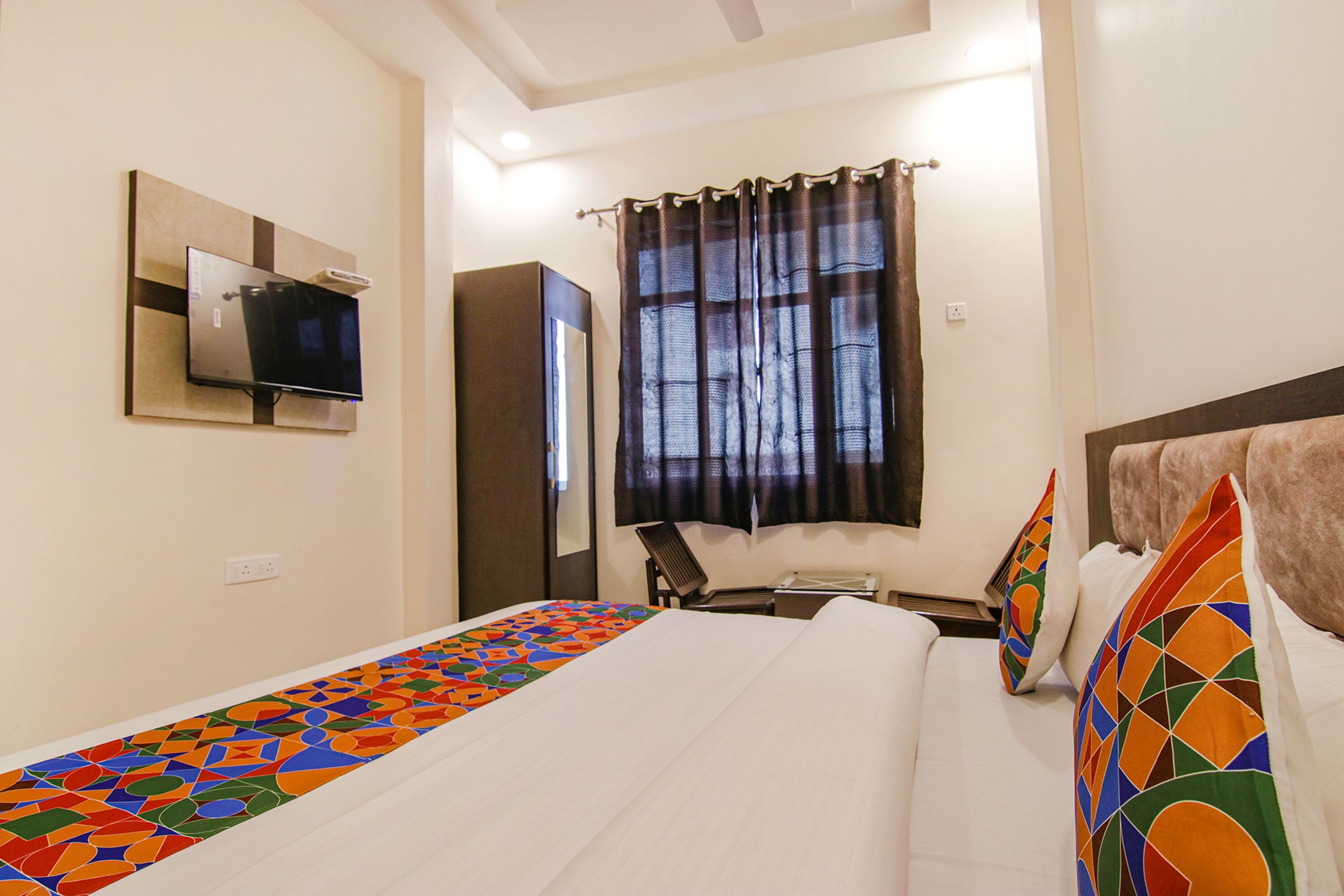 Quarto de hotel com cama, televisão na parede, escrivaninha e janela com cortinas.