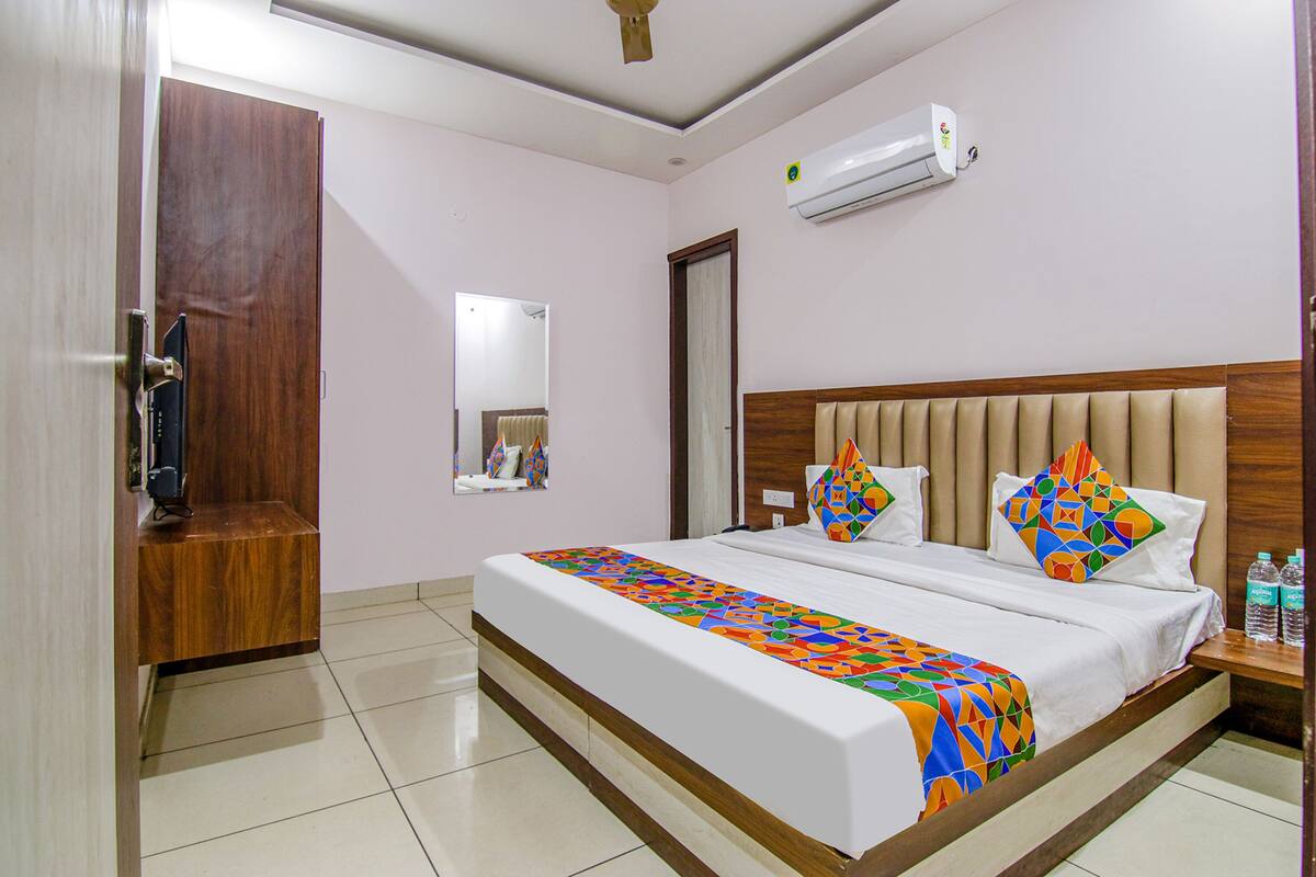 A hotel room with a bed, a wooden headboard, a bedside table with water bottles, a mirror, and an air conditioning unit.