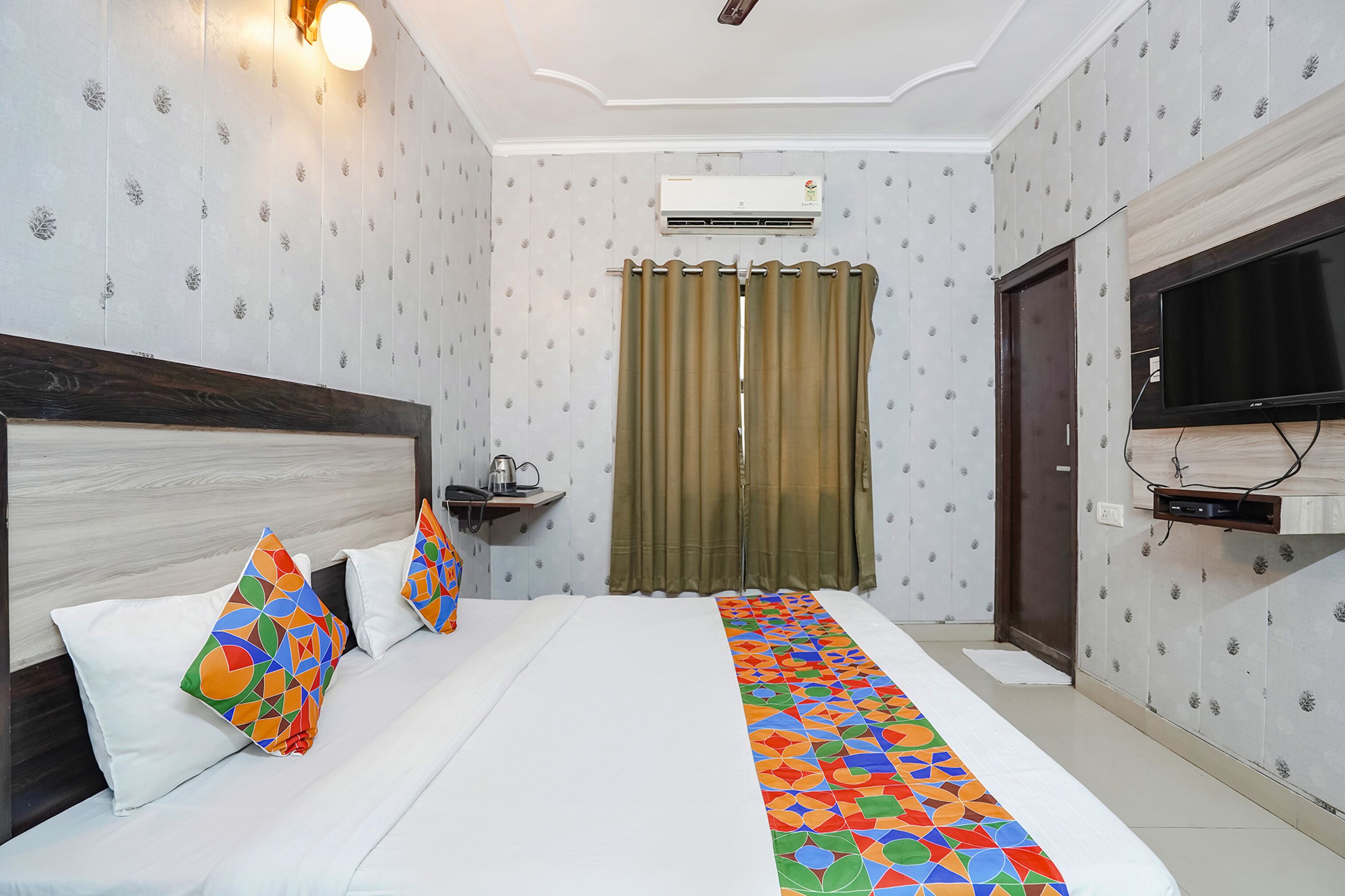 Habitación de hotel con cama, televisor, aire acondicionado instalado en la pared, cortina y una pequeña estantería con una tetera.
