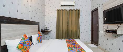 Habitación de hotel con cama, televisor, aire acondicionado instalado en la pared, cortina y una pequeña estantería con una tetera.