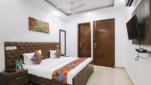 A bedroom with a tufted headboard, a bed with colorful pillows, a bedside table with water bottles, a ceiling fan, and a TV mounted on the wall. - FabExpress The Premium Villa (New Delhi)