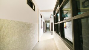 A corridor with a tiled floor, a white wall with a textured lower half, and a window with a view of a balcony. - Fabhotel Kempty Lake Resort (Dhanaulti)